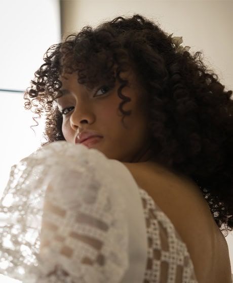 Una mujer con cabello rizado y un vestido de encaje blanco mira por encima del hombro hacia la cámara, con una suave luz natural iluminando un lado de su rostro.