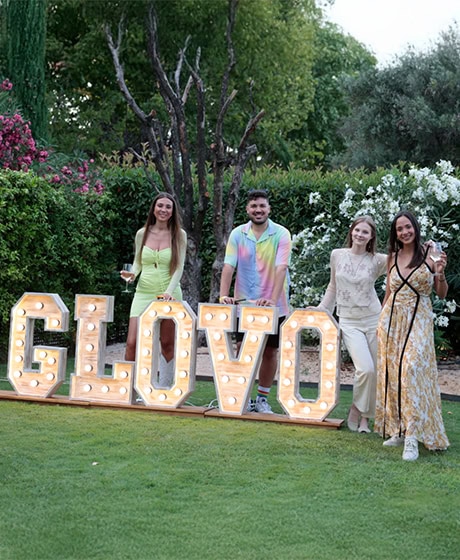 Cuatro personas están de pie sobre el césped junto a grandes letras iluminadas que forman la palabra "GLOVO", con árboles y arbustos en el fondo.