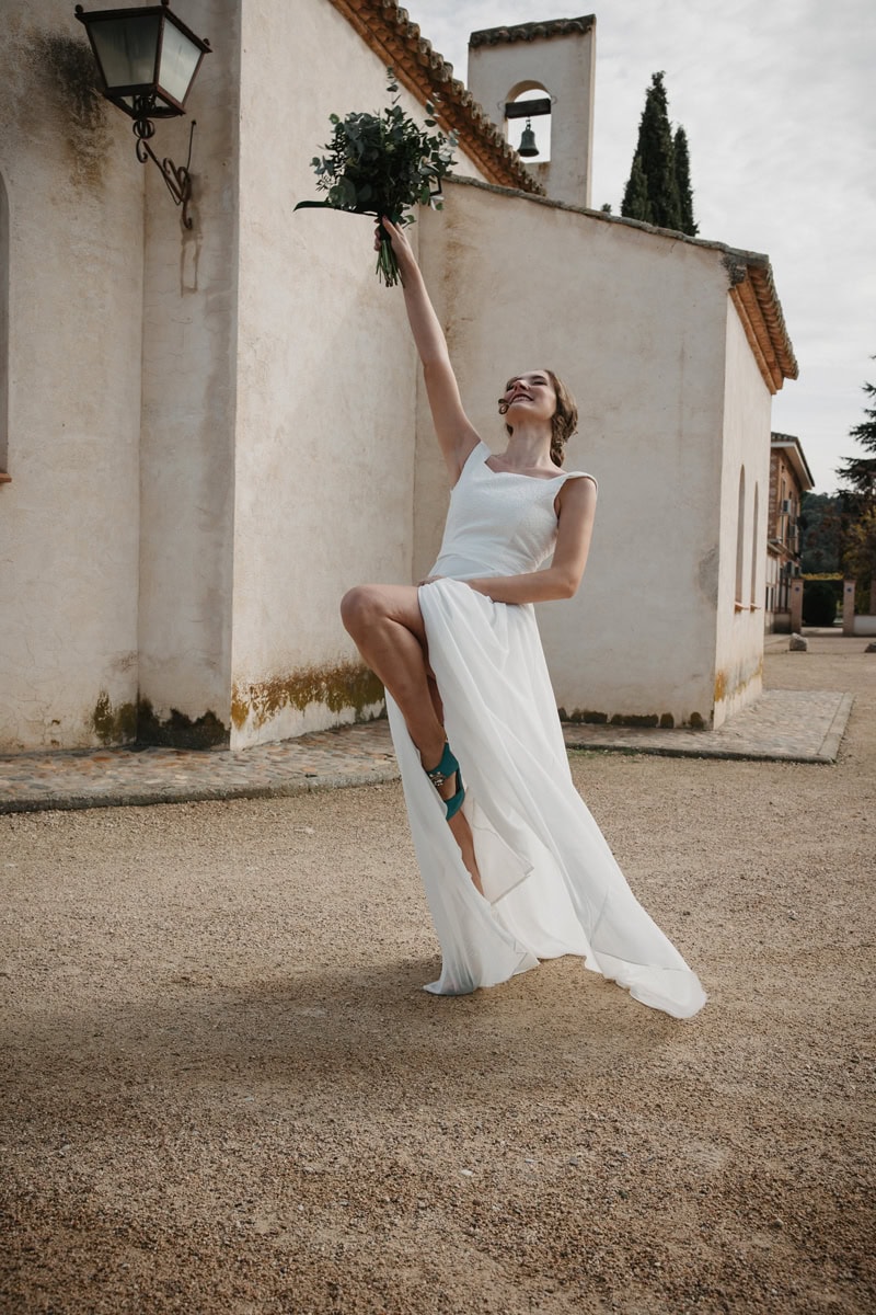 Una novia con un vestido blanco se encuentra al aire libre sobre una pierna, sosteniendo un ramo en alto y sonriendo alegremente frente a un edificio de color claro.