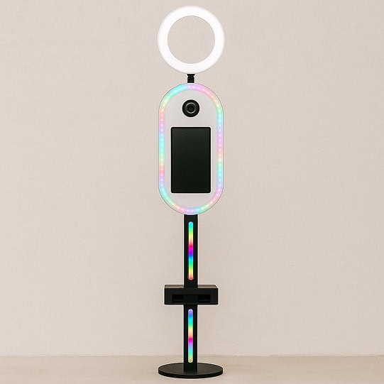Un fotomatón moderno con un anillo de luz en la parte superior, una cámara, una pantalla táctil y luces LED a lo largo de los bordes, todo montado en un soporte negro sobre un fondo liso.