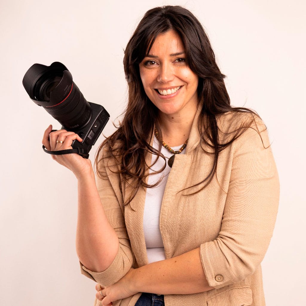 Nuria González, de cabello largo y oscuro, vestida con un blazer beige y una blusa blanca, sonríe mientras sostiene una cámara profesional contra un fondo claro.